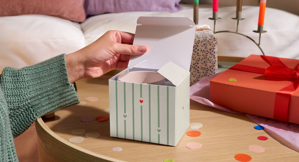 Person öffnet eine gestreifte Geschenkbox mit einer Tasse darin.