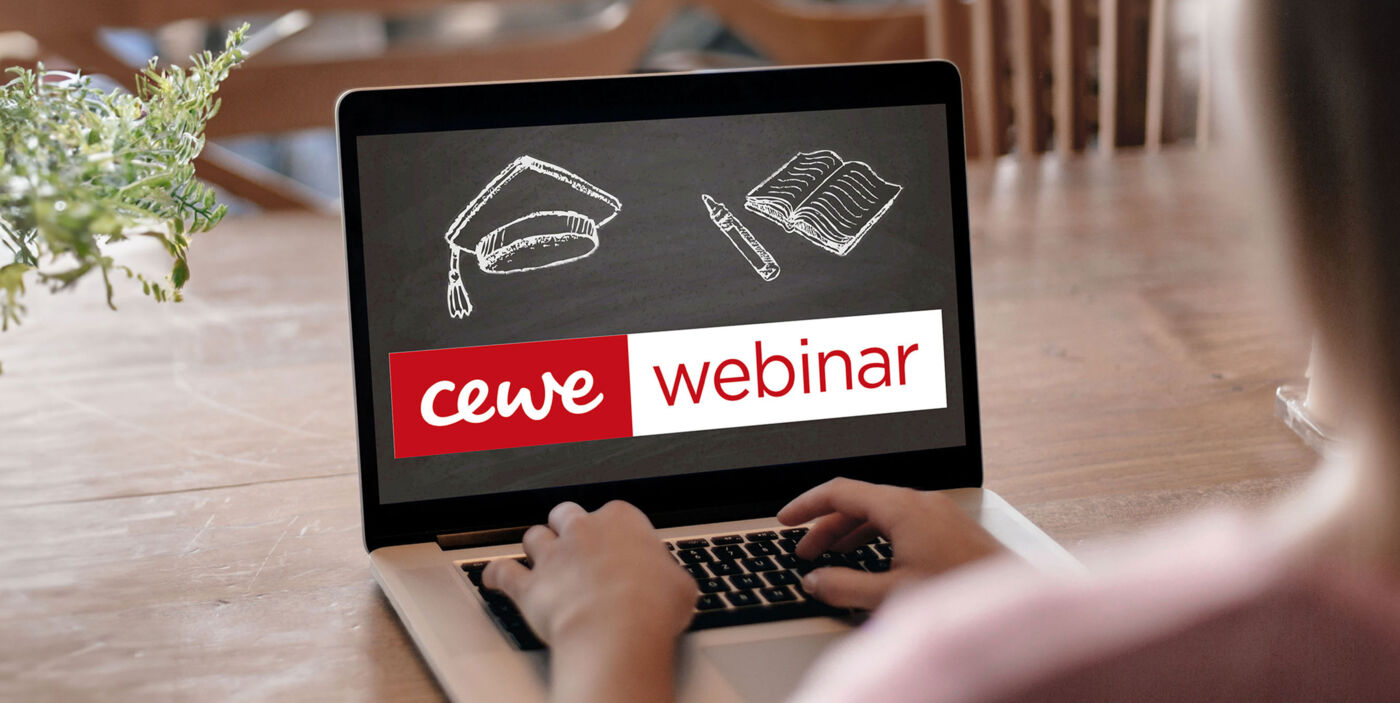 Eine Person sitzt an einem Tisch und arbeitet an einem Laptop. Auf dem Screen steht "CEWE Webinar".