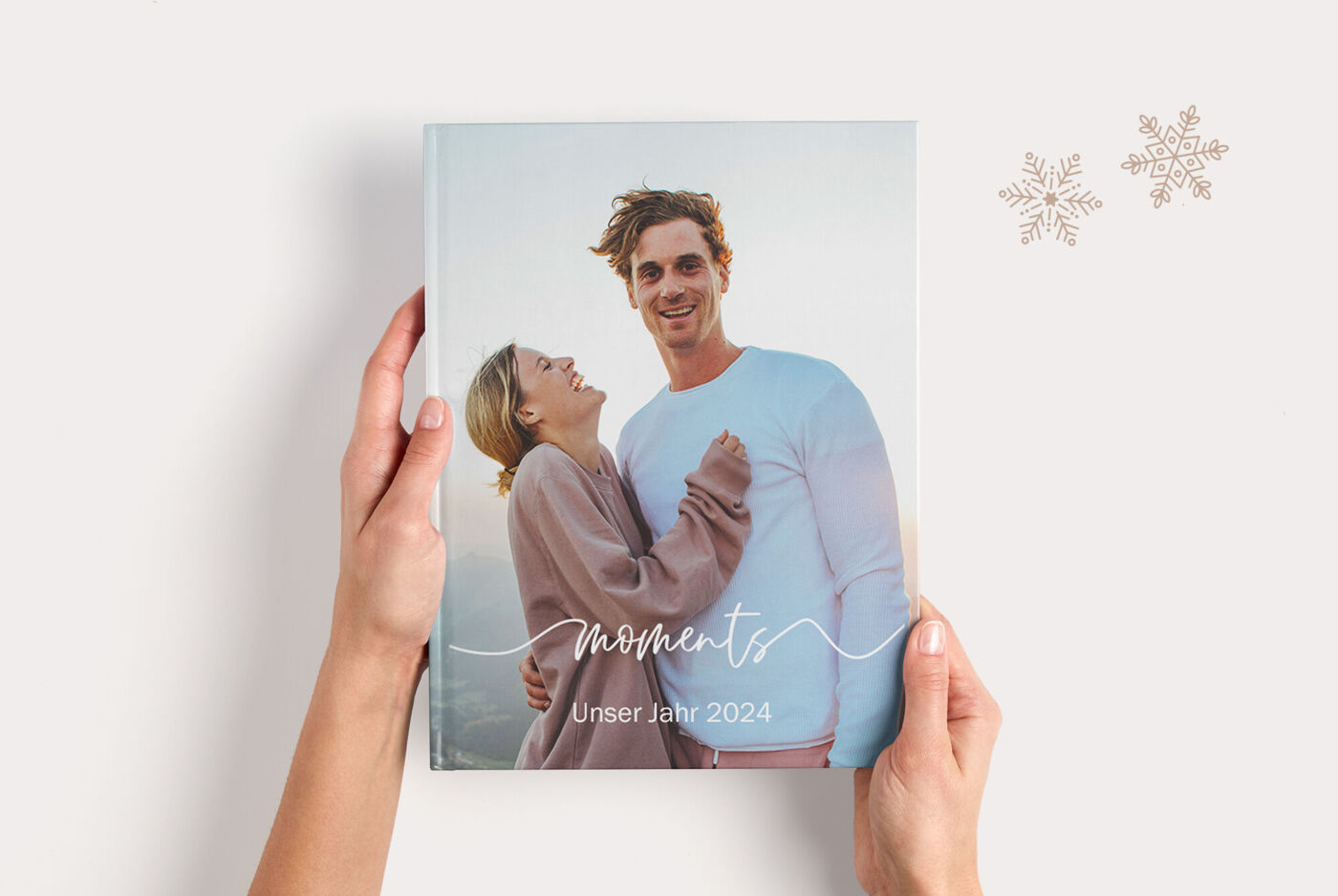 "Hände halten ein CEWE FOTOBUCH mit einem Paarfoto und dem Schriftzug „Moments Unser Jahr 2024“.         "